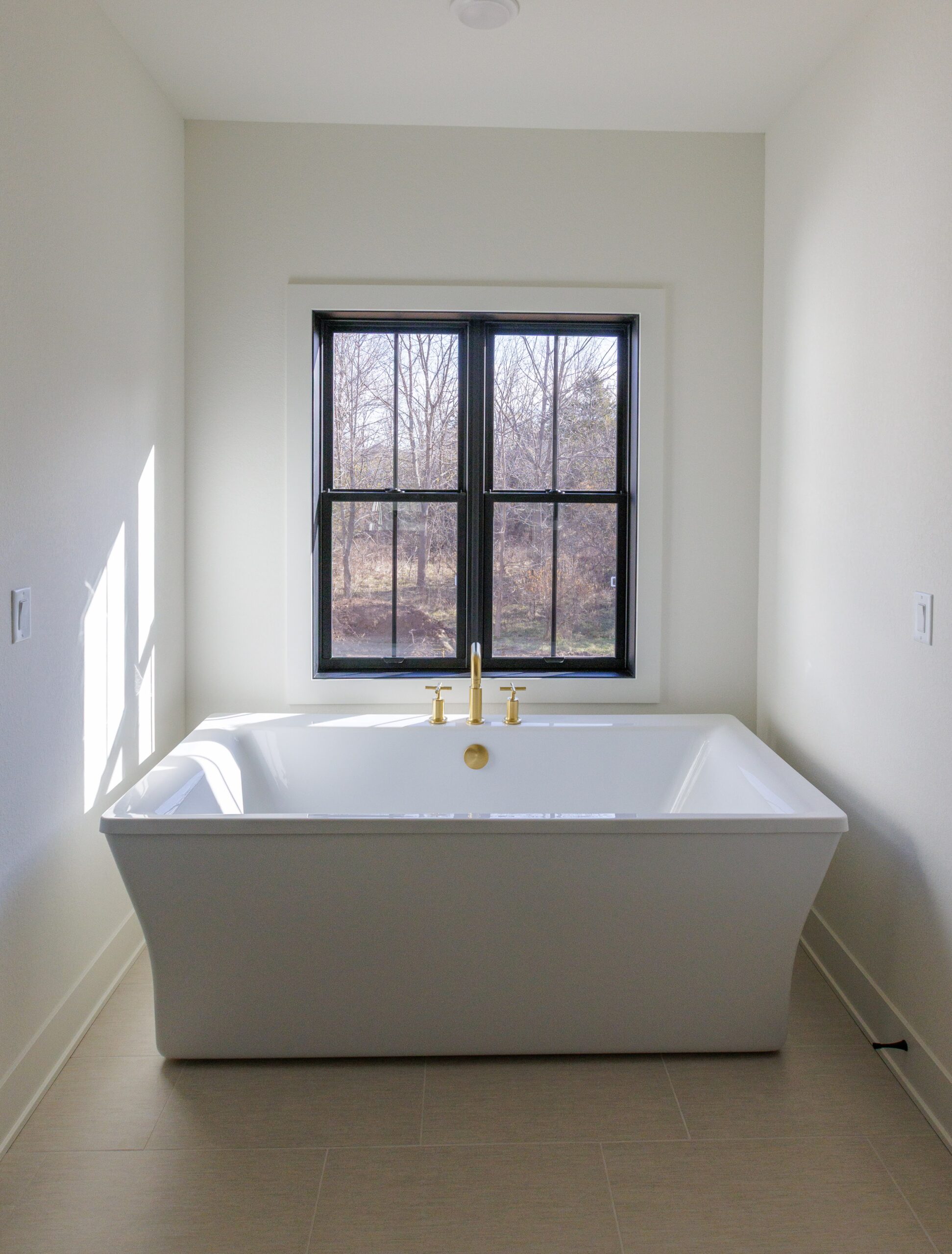 Freestanding white soaking tub with gold fixtures positioned beneath a window