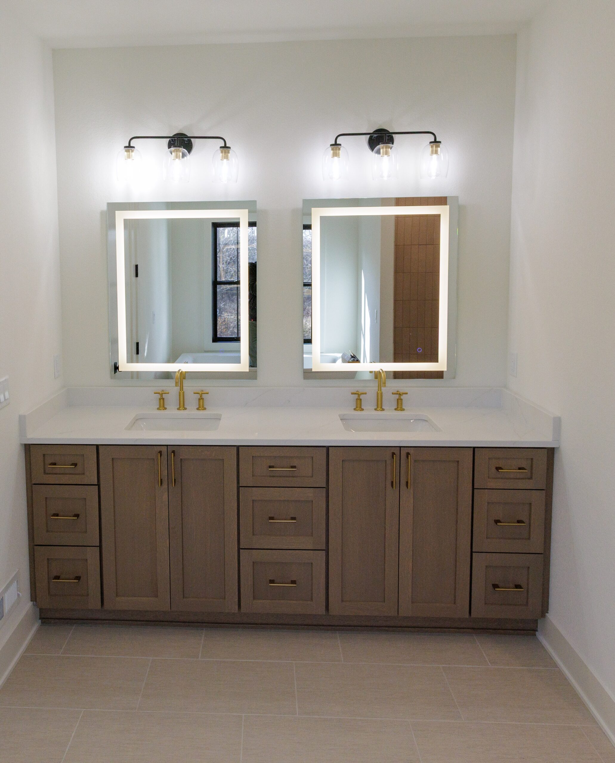 Bathroom with double vanity, backlit mirrors, wood cabinetry, and gold fixtures