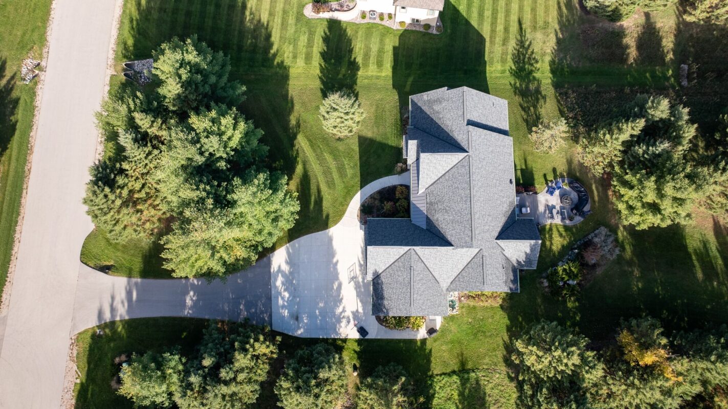 aerial view of custom home with curved driveway, landscaped yard, and surrounding trees